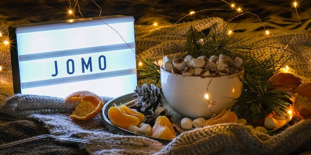 Lightbox with text JOMO Joy of missing out Cup with hot winter cacao and marshmallows tangerines spruce branch on bed. Christmas lights. Pine cones decoration. Cozy winter days. Hygge