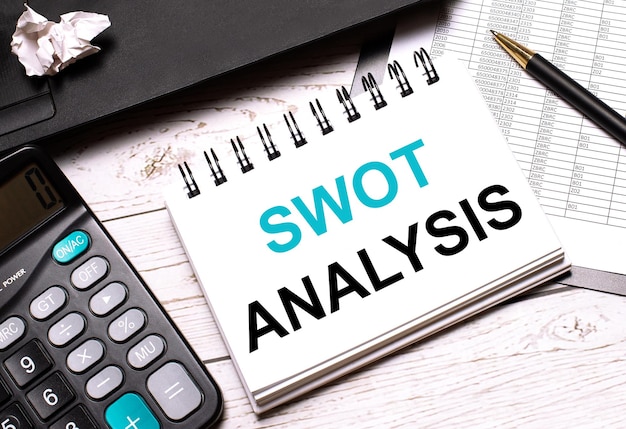 On a lightcolored office table is a computer a black calculator a pen and a notebook with the text SWOT ANALYSIS Business concept