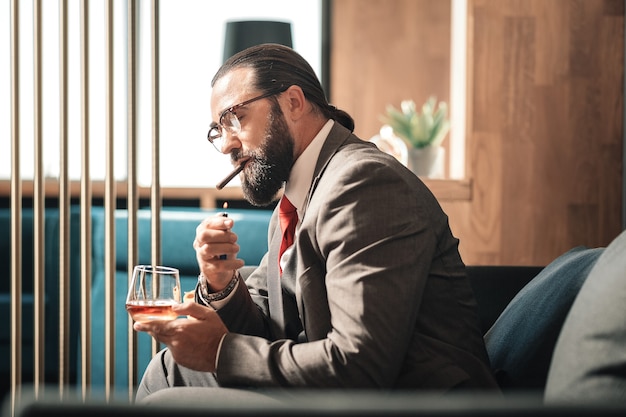 Lightning cigar. Bearded dark-haired man wearing glasses lightning his cigar while drinking whisky