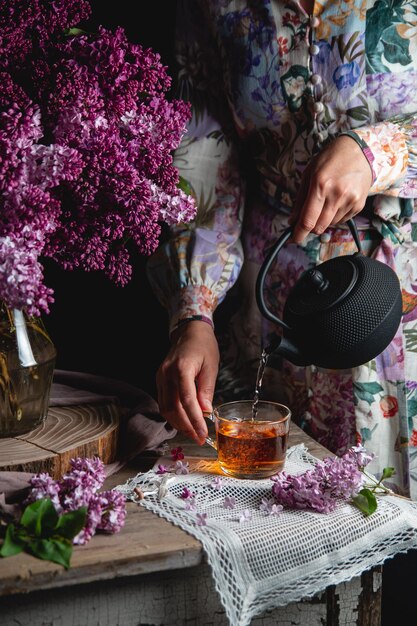 A lightskinned girl in a pastelcolored dress pours tea from a black castiron teapot Spring composition with lilacs on a vintage table Voluminous soft shadows Low key