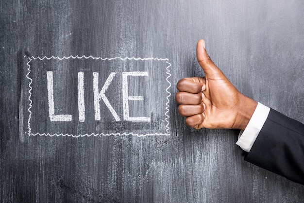 Like this! Close-up of human hand with thumb up in front of the blackboard with inscription Like