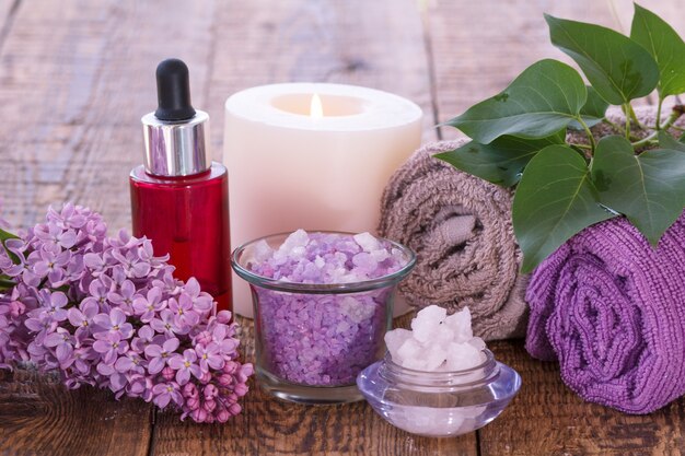 Lilac flowers, red bottle with aromatic oil, burning candle, bowls with sea salt and towels for bathroom procedures with green leaves on wooden boards. Spa products and accessories