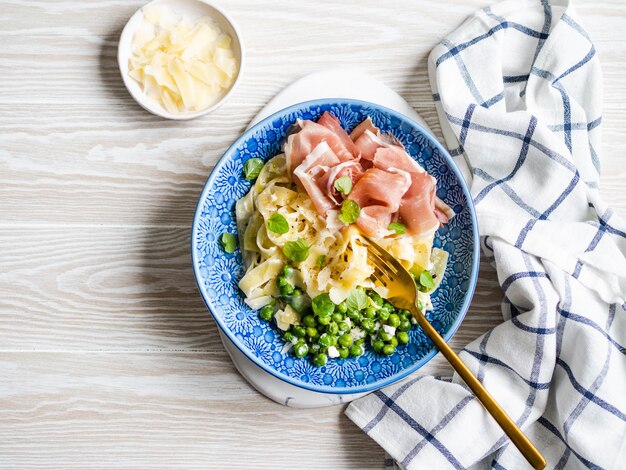 Linguine pasta with white sauce, green peas, Parmesan, prosciutto and fresh mint in a blue plate on wood board.