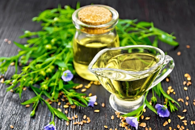 Linseed oil in a glass jar and gravy boat with seeds, leaves and flax flowers on a wooden board background