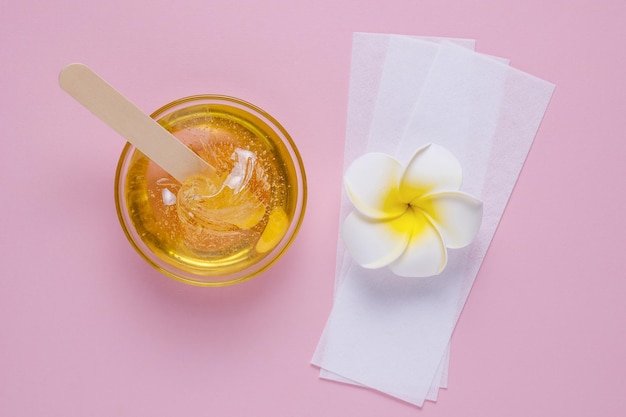 Liquid yellow sugar paste wooden spatula depilatory strips and flower on a pink background Sugaring