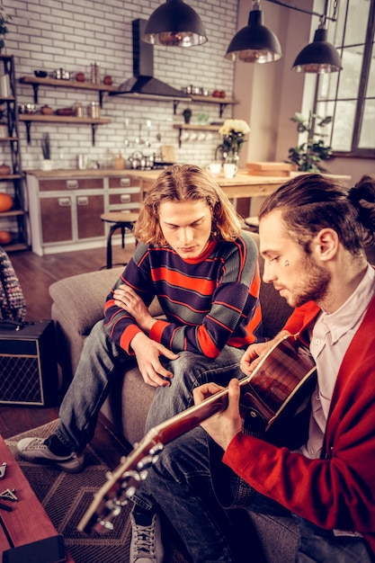 Listening to friend. Blonde-haired man wearing striped sweater listening to his friend playing the guitar