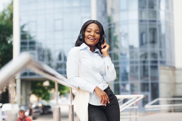 Listening to music in headphones Young afro american woman in white shirt outdoors in the city against business building