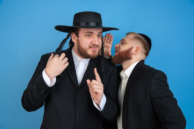 Listening to secrets. Portrait of a young orthodox jewish men isolated on blue wall. Purim, business, festival, holiday, celebration Pesach or Passover, judaism, religion concept.