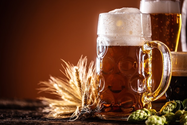 A liter glass full of draft beer next to it two smaller beers in front of a wooden barrel as a decoration of barley and hops.