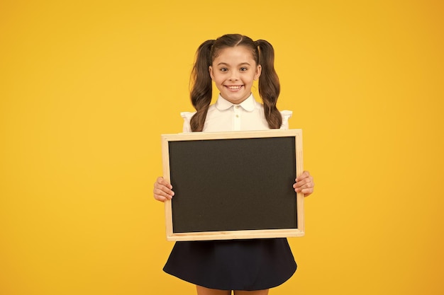 Little ad maker Little schoolchild holding blackboard on yellow background Little child with blackboard for school advertisement Little girl with empty chalkboard for your text copy space