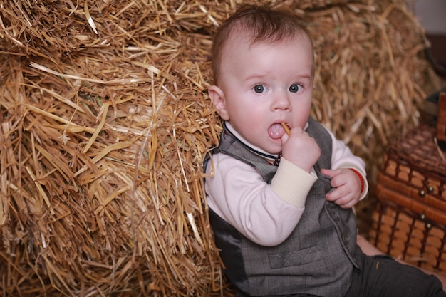 Little adorable baby boy sits near haystacks indoor and looks serious eyes.