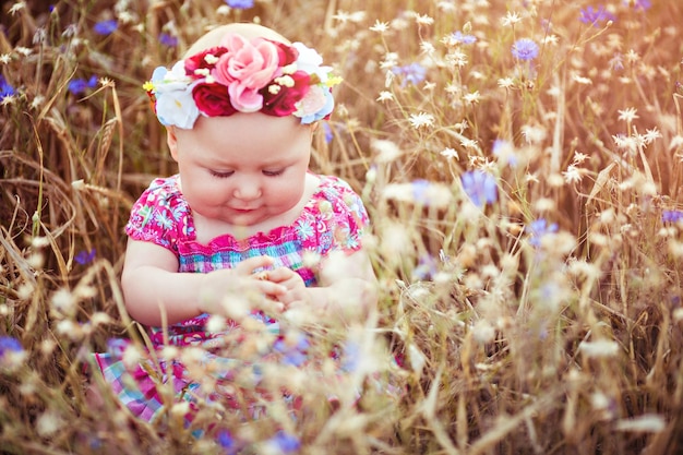 Little adorable baby girl with a flower wreath sitting on a summer flowering meadow