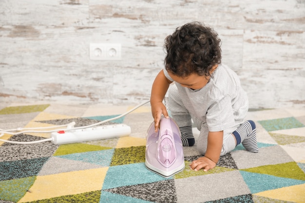 Little African-American baby playing with iron at home. Child in danger