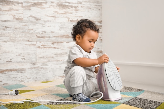 Little African-American baby playing with iron at home. Child in danger