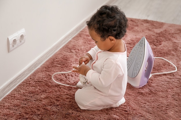 Little African-American baby playing with socket and iron at home. Child in danger