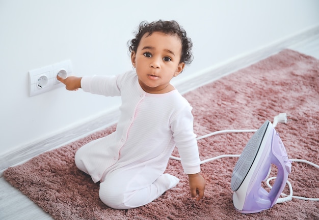 Little African-American baby playing with socket and iron at home. Child in danger