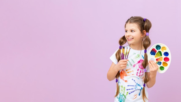 A little artist points to your advertisement on a pink isolated background The child holds a palette of colorful paints and brushes for drawing Art courses for schoolchildren