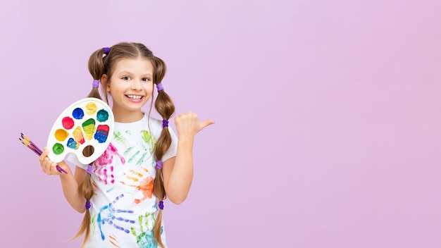 A little artist points to your advertisement on a pink isolated background The child holds a palette of colorful paints and brushes for drawing Art courses for schoolchildren