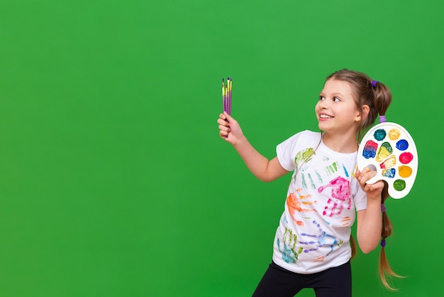 A little artist with a palette of colors points to your advertisement on a green isolated background Art courses for children