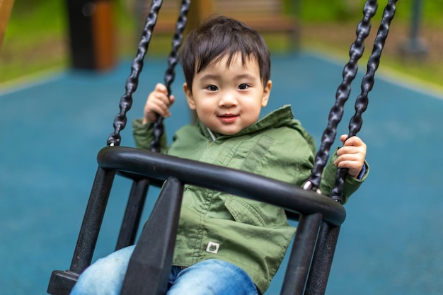 Little Asian boy swings on a swing on the wooden playground with happy face in summer outdoors and looking at the camera with a smile