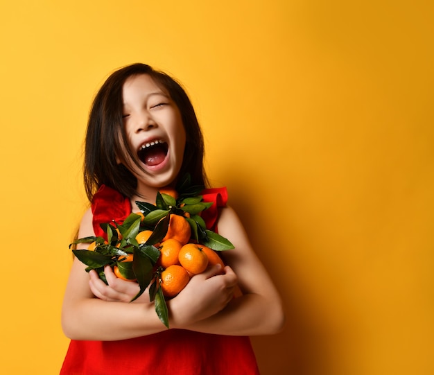 Little asian child in a red blouse. Laughing out loud with a toothless mouth, holding tangerines and oranges in their hands with green leaves, posing on an orange background. Childhood, emotions.