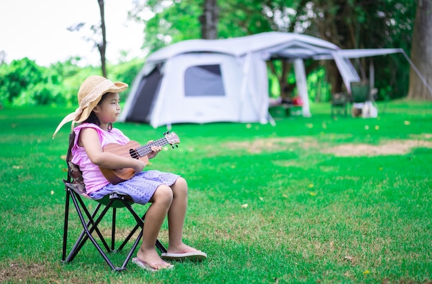 Little asian girl playing ukulele or hawaiian guitar in the park while camping in summertime