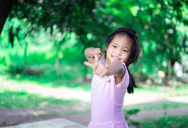 Little asian girl shooting wooden slingshot against green tree, active leisure for kids.