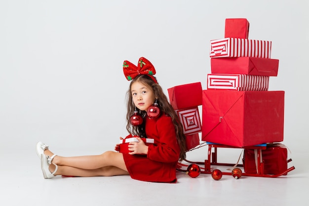 A little Asian girl sitting in a red dress sits with gift boxes on a white background. Christmas concept, text space