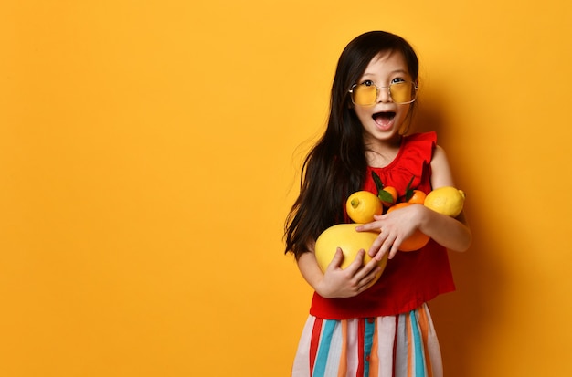 Little asian girl in sunglasses, brown beret, red blouse, colored skirt. Excited, holding pomelo, oranges and lemons in her hands, posing on orange background. Childhood, fruits. Close up, copy space