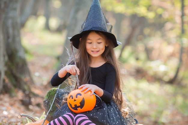 A little Asian girl in a witch costume for Halloween walks in the forest with a bucket for sweets