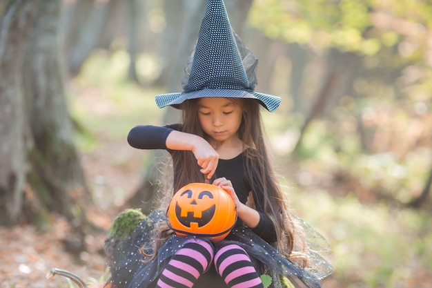 A little Asian girl in a witch costume for Halloween walks in the forest with a bucket for sweets