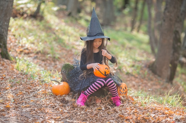 A little Asian girl in a witch costume for Halloween walks in the forest with a bucket for sweets