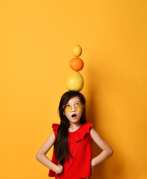 Little asian schoolgirl in sunglasses, red blouse, colored skirt. Amazed, holding pomelo, orange and lemon on her head, posing on orange background. Childhood, fruits, emotions. Close up, copy space