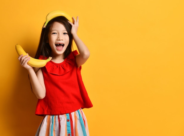 Little asian woman in red blouse, colorful skirt. Looking surprised, holding one banana in his hand, the other lay on his head, toothless mouth, posing against an orange background. fruits, emotions