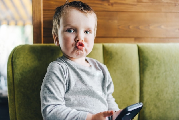 Little baby boy sitting on sofa with mobile phone in hands and sending a kiss