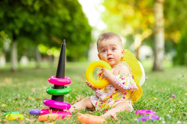 Little baby girl 7 months old sitting on the green grass in a yellow dress and hat and playing with a pyramid