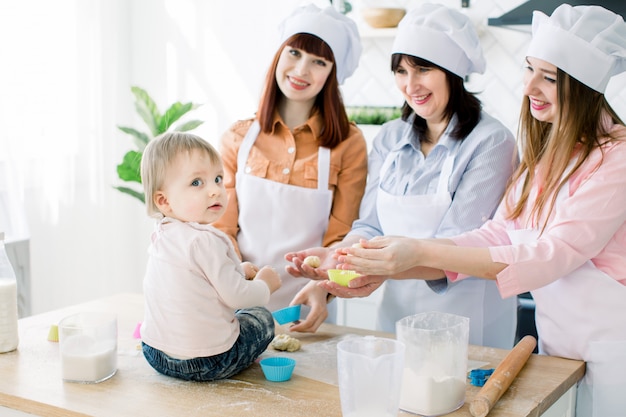 Little baby girl helps to make cupcakes together with mother, aunt and grandmother. Pretty women are smiling and having fun while baking together at home kitchen