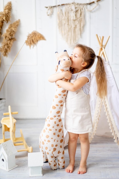 A little baby girl is playing with a big giraffe as tall as she is at home in a bright nursery and laughing