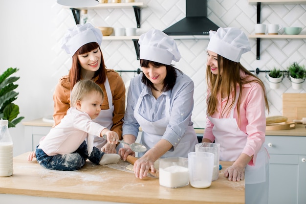 Little baby girl is sitting on the wooden table at kitchen while her mother, aunt and grandmother are making the dough for cookies. Happy women in white aprons baking together