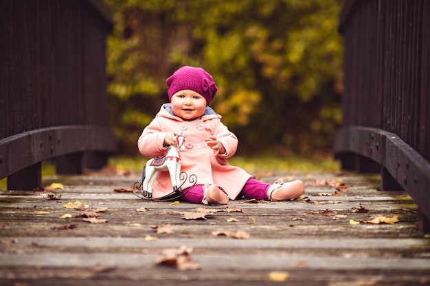 Little baby girl in knitted hat and jacket sitting on the wooden bridge with a lot of autumn leaves in park