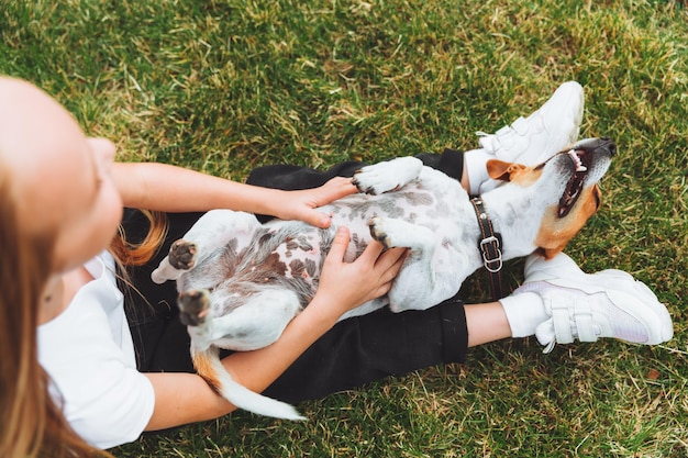 A little baby girl scratches the belly of her Jack Russell Terrier dog A happy dog is lying on the green grass in the park a pet