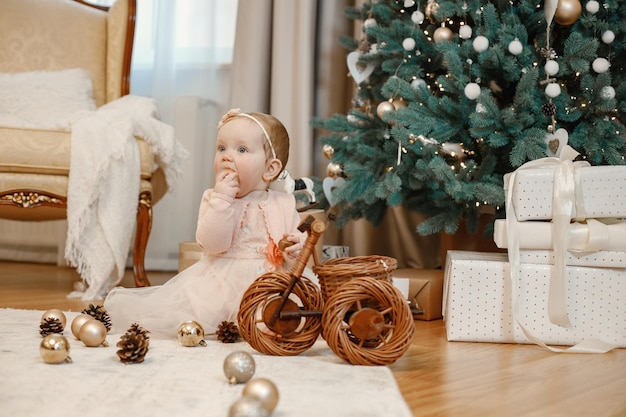 Little baby girl sitting on the floor near christmas tree at home. Girl is playing with balls for Christmas tree decoration. Girl wearing peach dress.