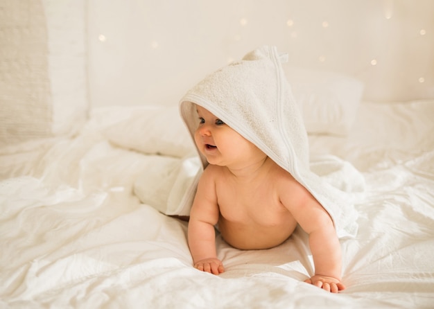 Little baby girl in a white towel with a hood is onll fours on white blanket on the bed