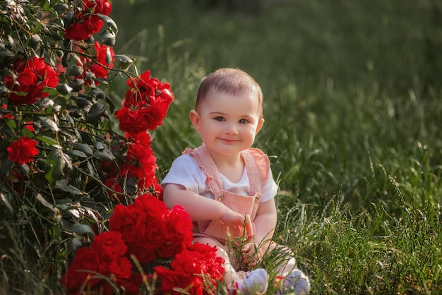 A little baby sits on a green lawn next to red roses. A happy girl in a powdery overalls enjoys a walk in the park. Closeup portrait of a child. Carefree childhood