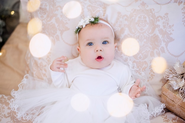 A little baby in a white dress lies on a light sofa in garlands of lights, a festive Christmas tree