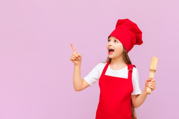 A little baker girl in a chef's hat and apron holds a rolling pin and points at your advertisement on a pink isolated background Copy space