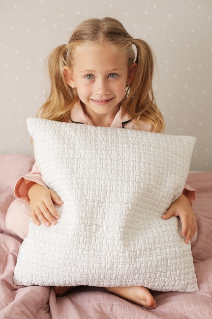 little beautiful girl in pink pajamas sits on a pink blanket on the bed at home hugs a pillow