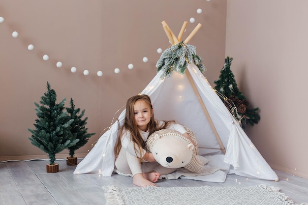 Little beautiful girl in white pajamas plays with a teddy bear near the teepee childrens room with c