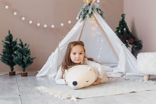 Little beautiful girl in white pajamas plays with a teddy bear near the teepee childrens room with c