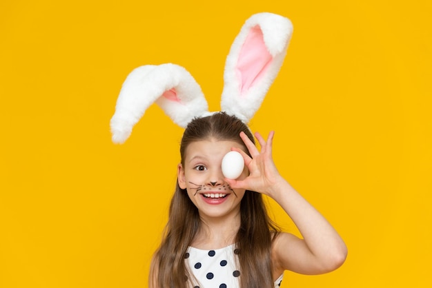 A little beautiful girl with masquerade ears of an Easter bunny on her head holds a chicken Easter egg in her hands and enjoys the Easter feast on a yellow isolated background